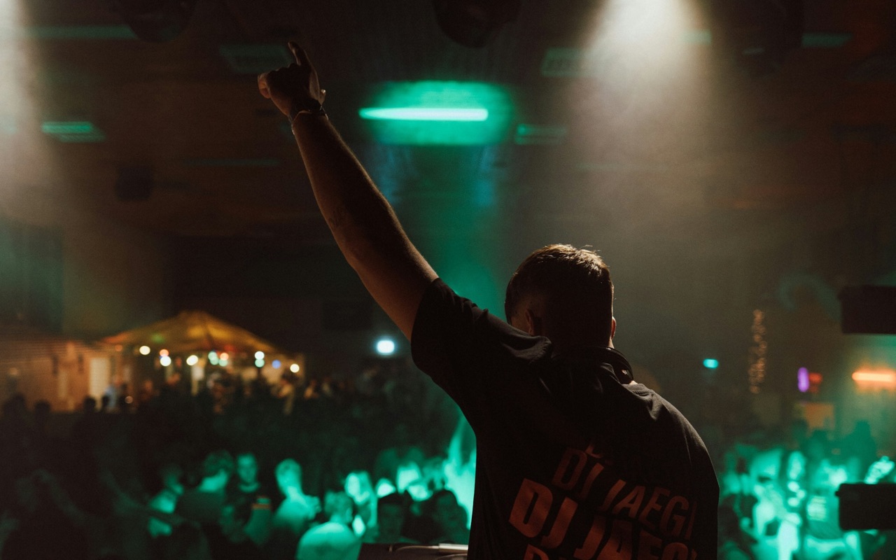 A DJ with his arm raised over a packed, green-lit dancefloor